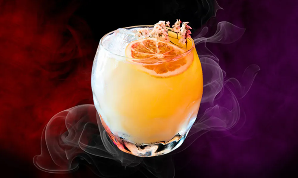 Short tumbler with gradient orange-yellow cocktail and ice, garnished with orange slice and dried flowers, red-purple smoky background