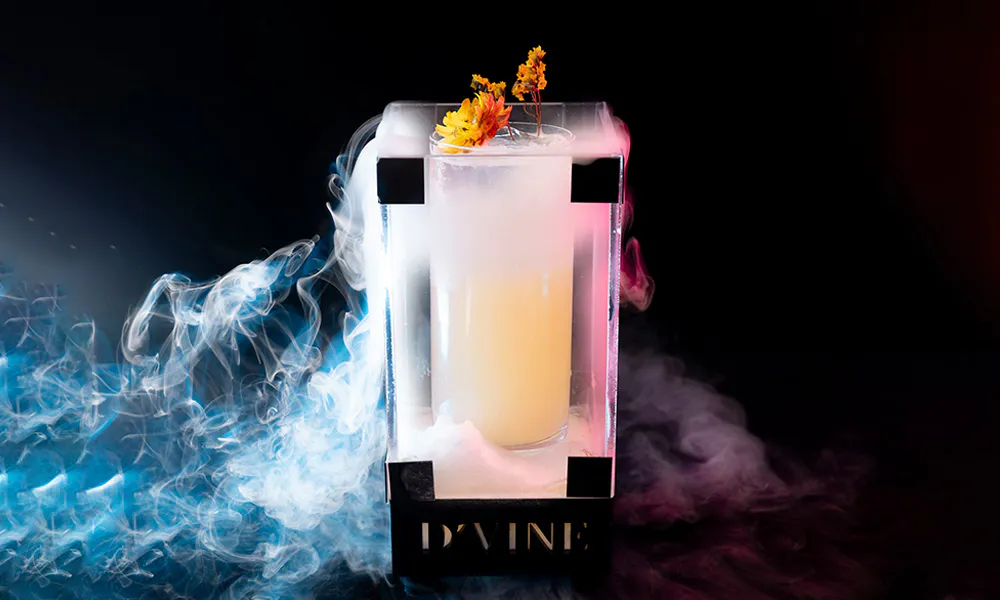 Pale cocktail in D'VINE acrylic box, dry ice creates blue and pink smoke, garnished with yellow dried flowers.