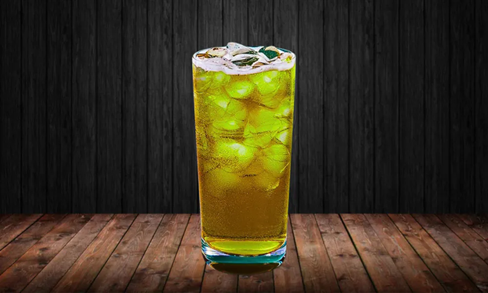 Tall clear glass filled with bubbly yellow-green beverage and ice cubes, on wooden table against dark wood panel background.