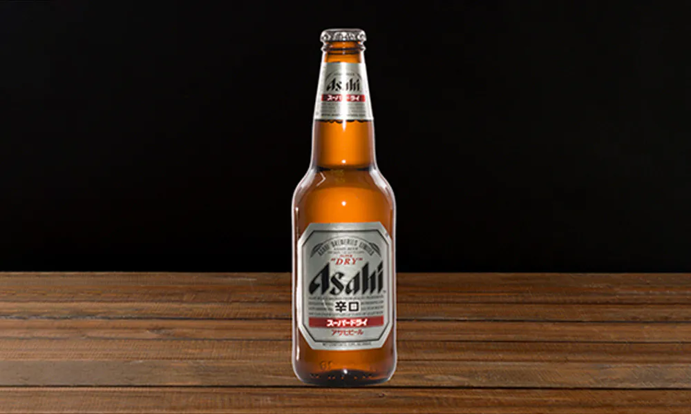 Brown glass Asahi beer bottle with silver label marked Asahi Super Dry, placed on wooden planks with dark background.