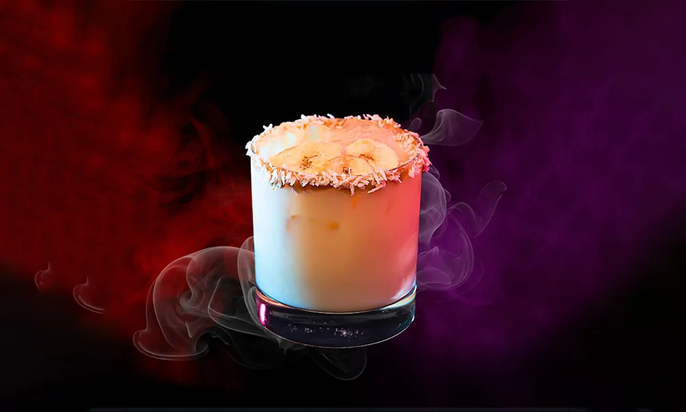 Low rocks glass with coconut rim, creamy drink topped with banana slices, surrounded by red and purple smoke on black background.